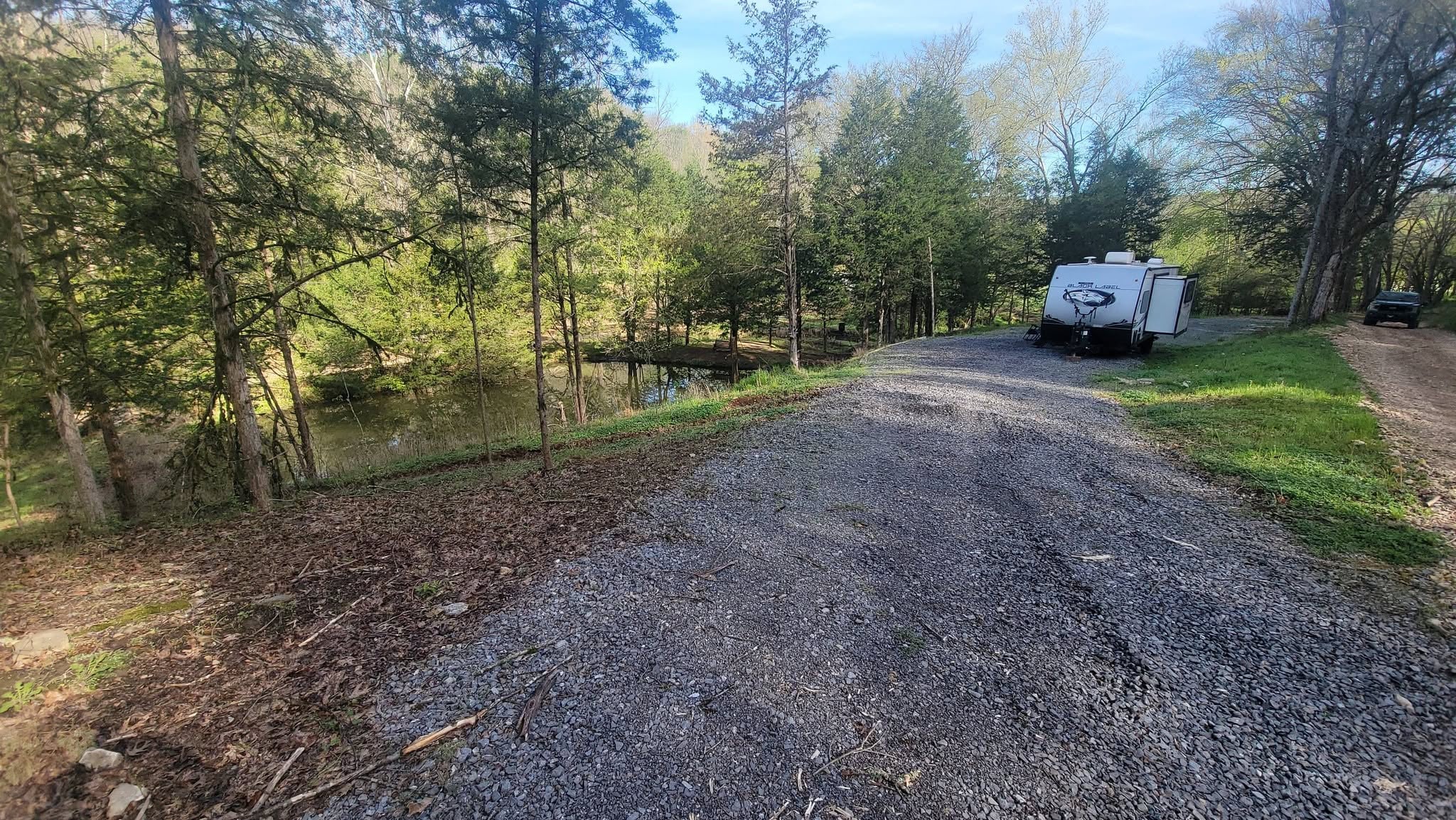 A white travel trailer is parked on a gravel road near a pond, surrounded by trees and greenery under a clear sky.