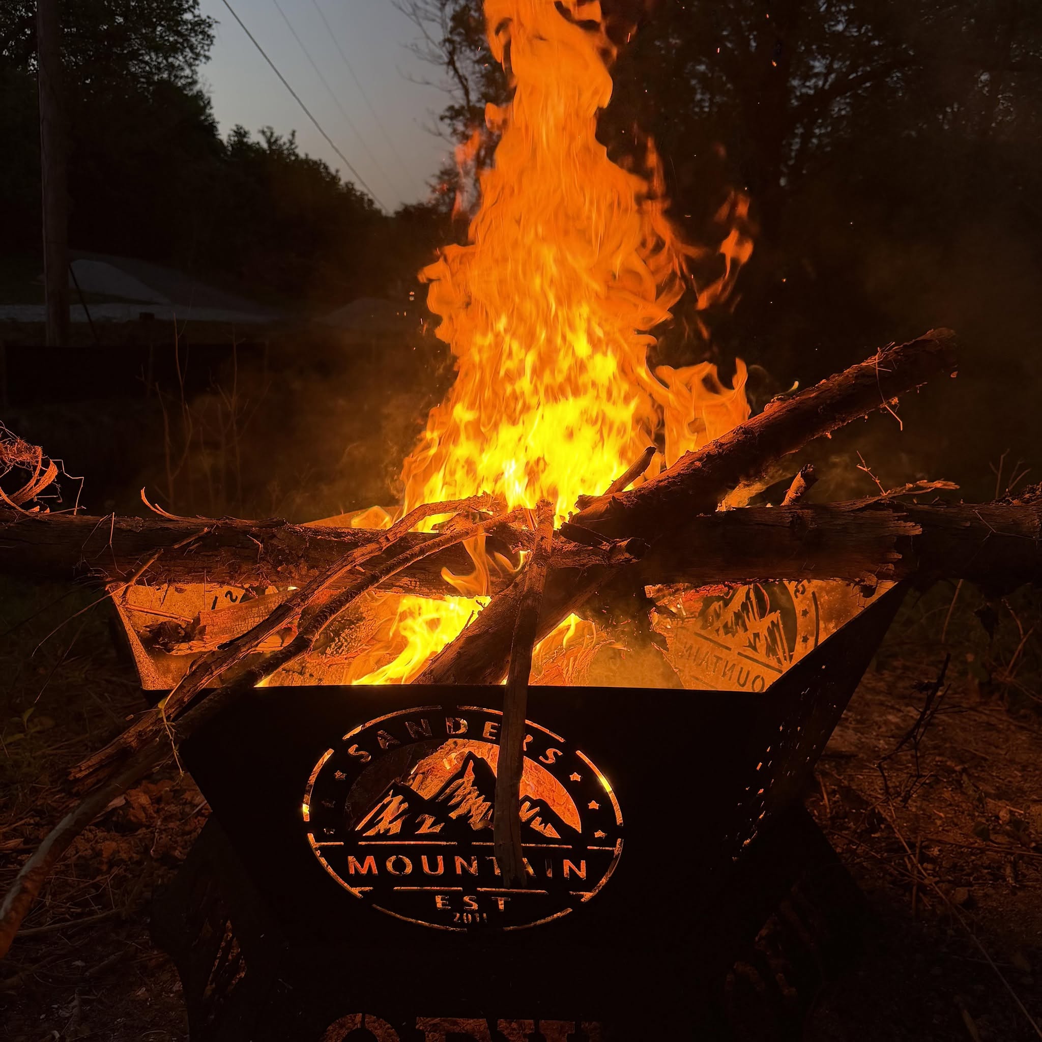 A campfire burns brightly in a metal fire pit with “Sanders Mountain” and a mountain graphic cut into the side. Logs rest across the top, and trees are visible in the background at dusk.