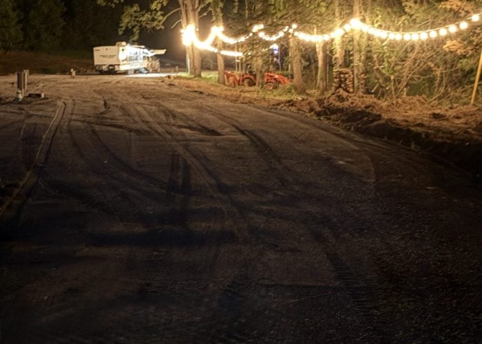 A dirt road at night lit by string lights, with trees on one side and a trailer in the background.