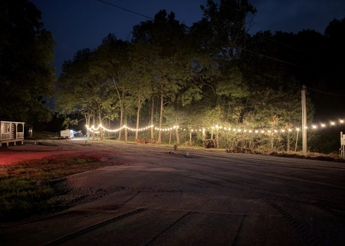 String lights are hung along a dirt road at night, illuminating trees and the road surface.