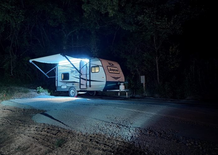 A small travel trailer with an extended awning is parked on gravel at night, illuminated by bright blue lights, with trees in the background.
