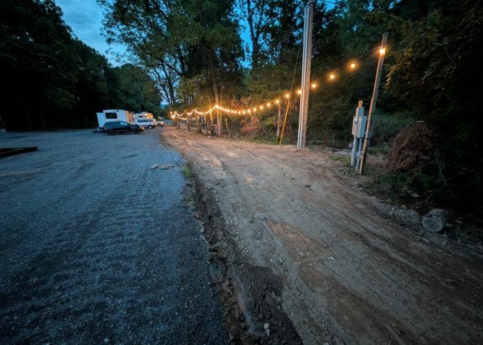 String lights hang above a dirt path next to gravel and parked vehicles, bordered by trees at dusk.