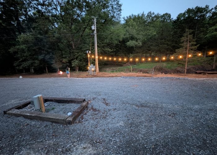 A gravel lot with a small wooden planter in the foreground and string lights hanging across trees in the background. Two people stand near a small campfire.