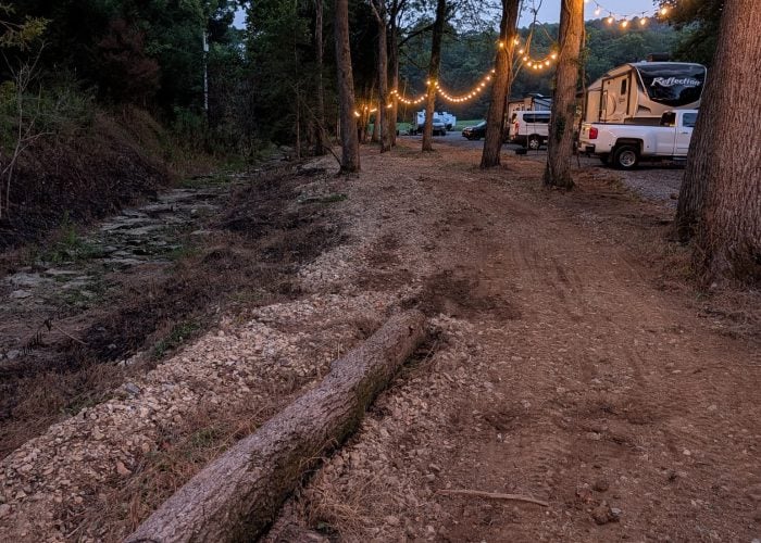 A dirt path with a fallen log leads to RVs and string lights hanging between trees at a campsite.