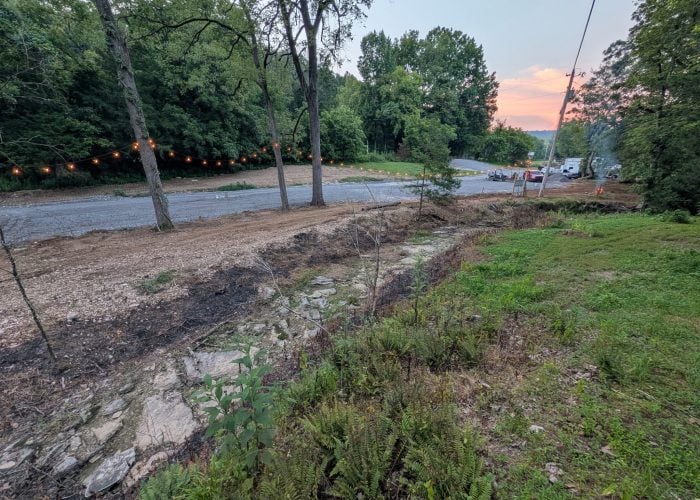 A gravel road runs through a wooded area at dusk, with string lights on trees and traffic cones near parked vehicles in the background.